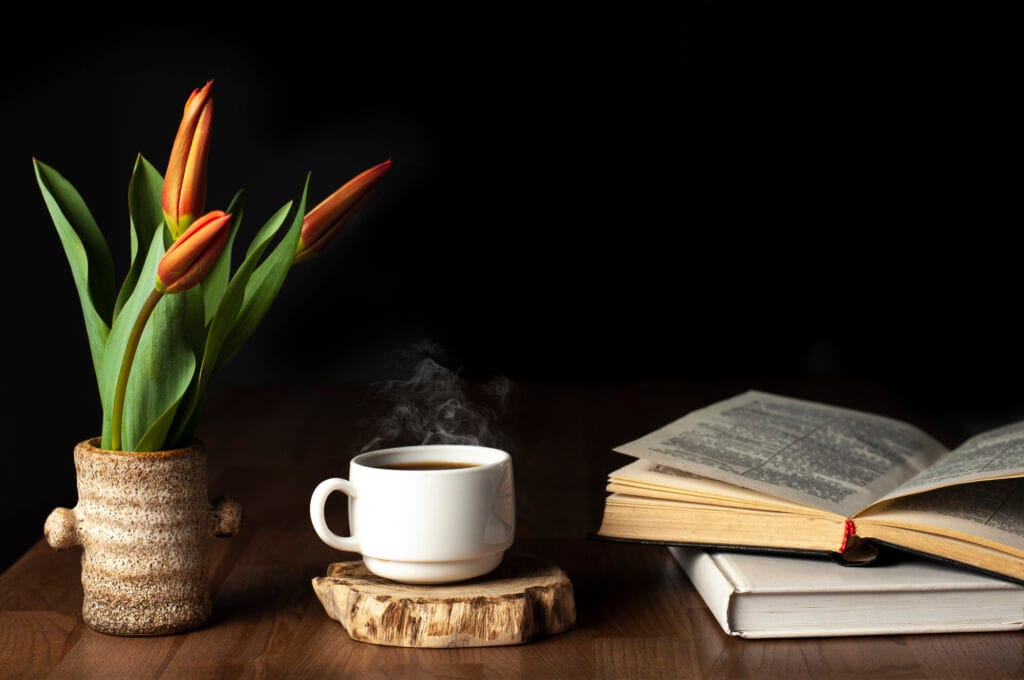 A centered cup of steaming coffee sets between an pottery vase filled with emerging bloom tulips and two books with the top book laying open to the center.