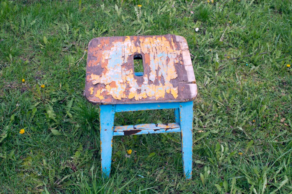 A distressed square topped white and blue wooden stool with carrying hole in center of the seat sets on a green lawn.