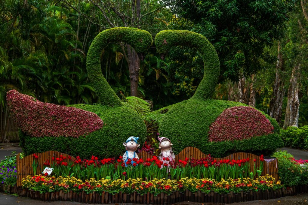 A boy and girl garden gnomes, surrounded by blooming spring flowers, look skyward while stnading in front of creative swan-shaped topiary that forms a heart shaped arch.